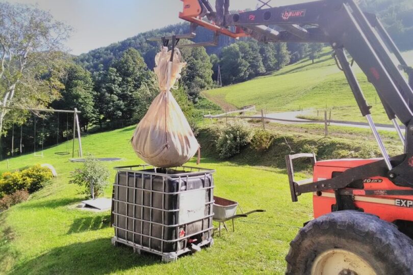 Ein Traktor mit Frontlader hebt einen großen, in einem Tuch verpackten Ballen über einen IBC-Tank. Die Szene spielt sich auf einer Wiese in hügeliger Landschaft ab. Im Hintergrund sind Bäume, ein Spielplatz und ein Feldweg sichtbar.