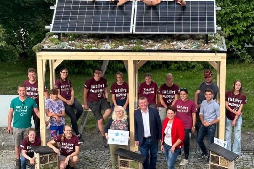 Gruppe von Jugendlichen und Erwachsenen posiert vor einem hölzernen Unterstand mit Solarpaneldach. Einige stehen darauf, andere darunter. Umgeben von Insektenhotels und bunten Boxen, scheint es sich um ein gemeinschaftliches Umwelt- oder Jugendprojekt zu handeln.
