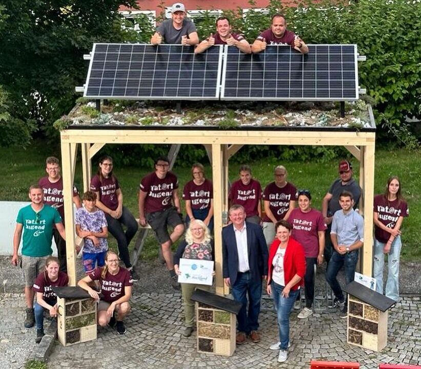 Gruppe von Jugendlichen und Erwachsenen posiert vor einem hölzernen Unterstand mit Solarpaneldach. Einige stehen darauf, andere darunter. Umgeben von Insektenhotels und bunten Boxen, scheint es sich um ein gemeinschaftliches Umwelt- oder Jugendprojekt zu handeln.