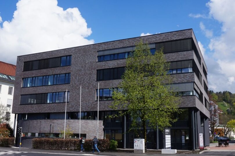 Modernes Bürogebäude mit dunkler Klinkerfassade und großflächigen Fenstern, fünf Geschosse, vorgelagerter Baum und Gehweg, blauer Himmel mit Wolken, angrenzende Straße mit Zebrastreifen im Vordergrund.