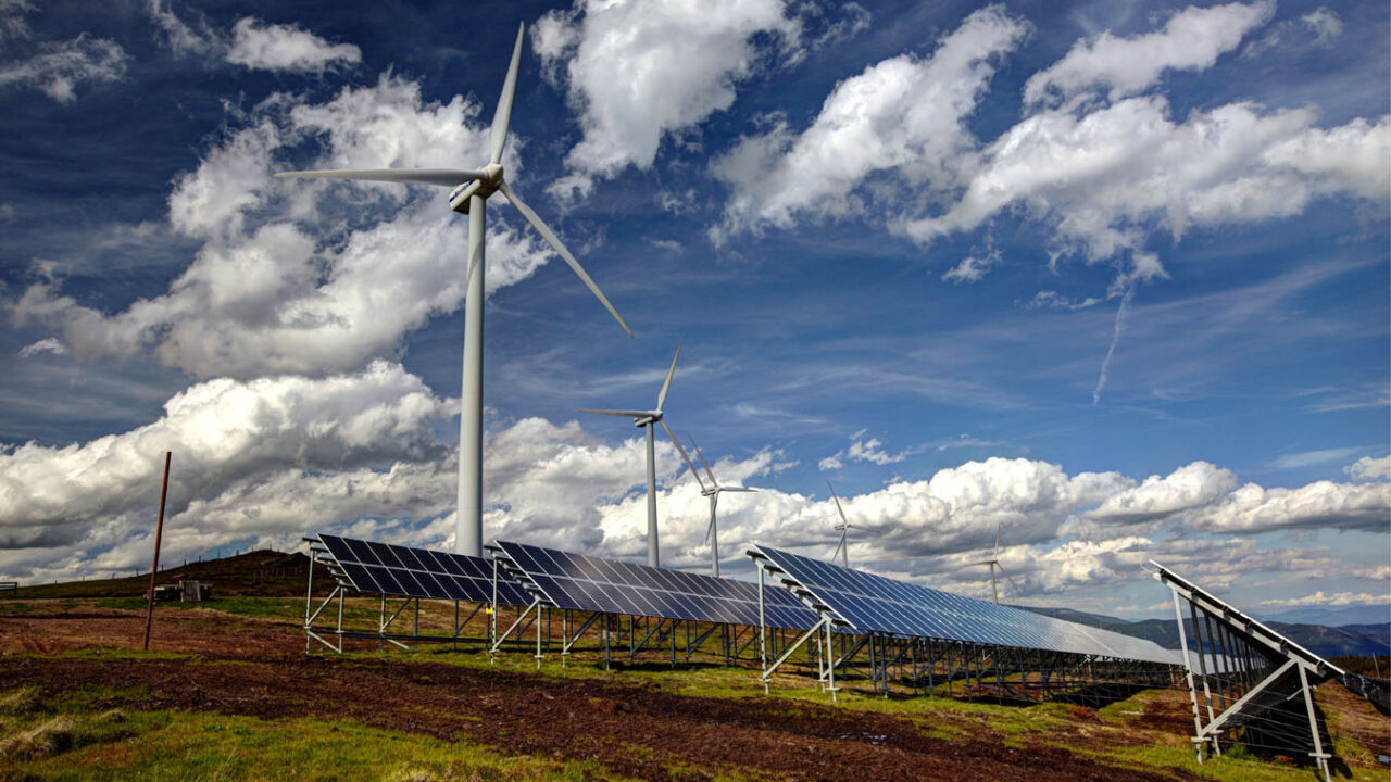 Mehrere Windräder und Solarpaneele stehen auf einer hügeligen Landschaft unter einem teils bewölkten Himmel. Die Kombination von Wind- und Solarenergieanlagen symbolisiert nachhaltige Stromerzeugung aus erneuerbaren Quellen.