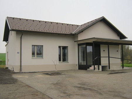 Eingeschossiges Gebäude mit hellem Putz, Satteldach und barrierefreiem Eingang mit Rampe. Asphaltierte Fläche davor, im Hintergrund Felder und Bäume. Mögliche Nutzung als Praxis oder Büro.