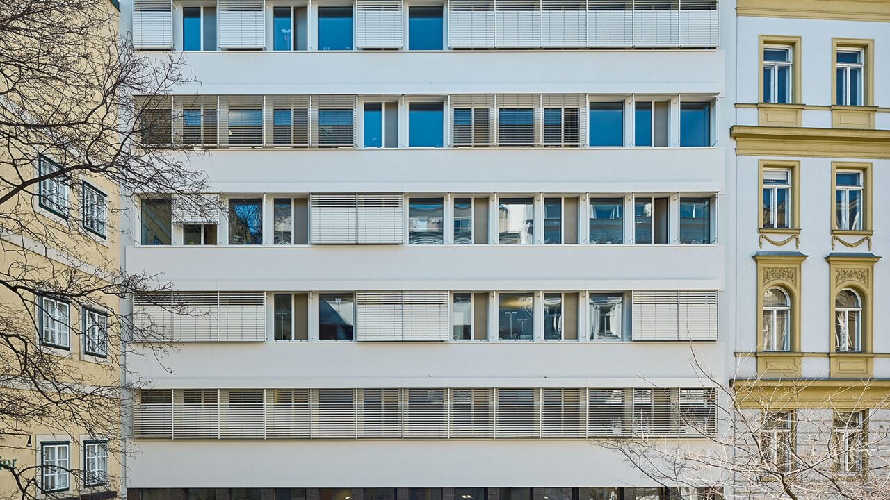 Frontalansicht eines modernisierten Bürogebäudes mit weißen Fassaden und horizontalen Sonnenschutzlamellen. Das Gebäude ist von zwei Altbauten eingerahmt und steht hinter einer begrünten Fläche mit Laterne.