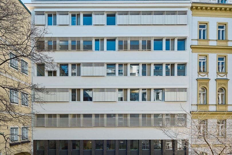 Frontalansicht eines modernisierten Bürogebäudes mit weißen Fassaden und horizontalen Sonnenschutzlamellen. Das Gebäude ist von zwei Altbauten eingerahmt und steht hinter einer begrünten Fläche mit Laterne.