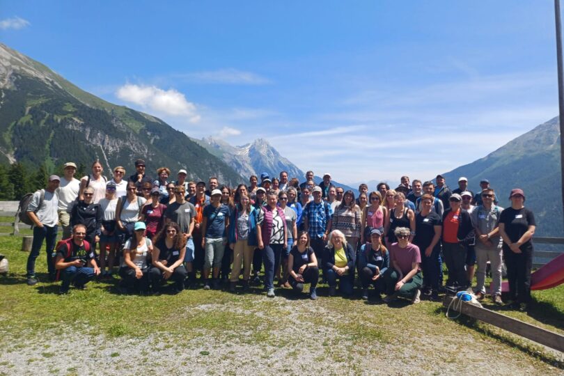 Große Gruppe von Menschen posiert auf einer Wiese vor Berglandschaft mit blauem Himmel und Wolken