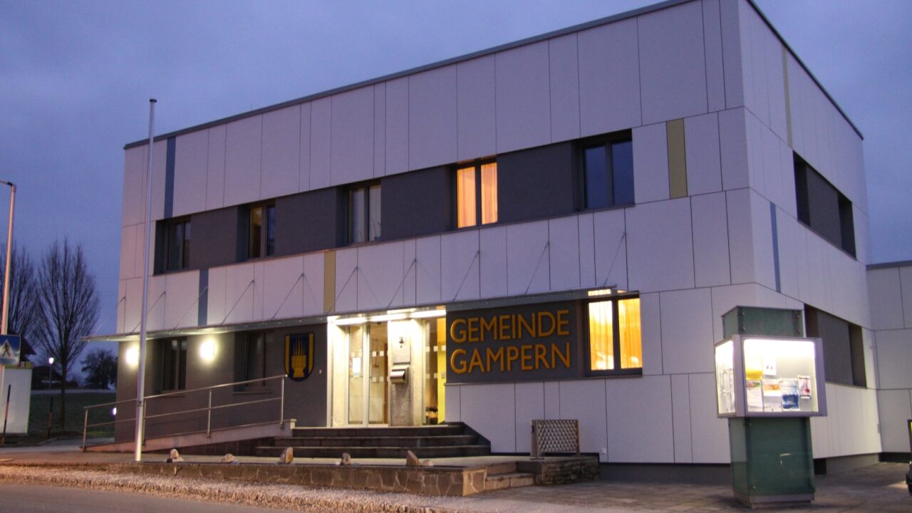 Modernes Gemeindeamt Gampern bei Abenddämmerung, beleuchtet von warmem Licht, mit klaren architektonischen Linien und großen Fenstern, davor eine Informationstafel, der Himmel ist bewölkt.