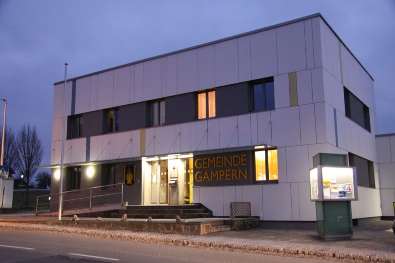 Modernes Gemeindeamt Gampern bei Abenddämmerung, beleuchtet von warmem Licht, mit klaren architektonischen Linien und großen Fenstern, davor eine Informationstafel, der Himmel ist bewölkt.
