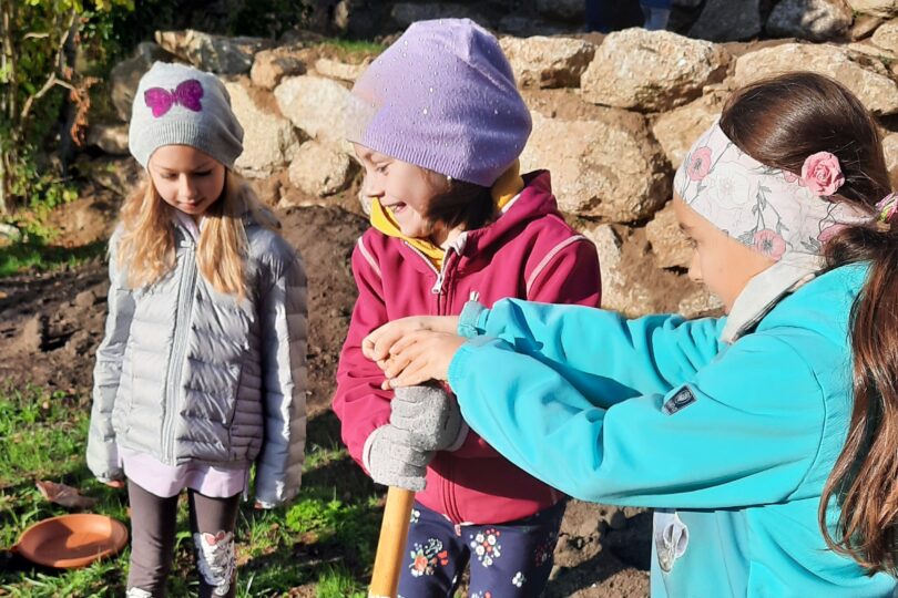 Drei Kinder stehen in Winterkleidung auf einer Wiese und buddeln mit einer Schaufel