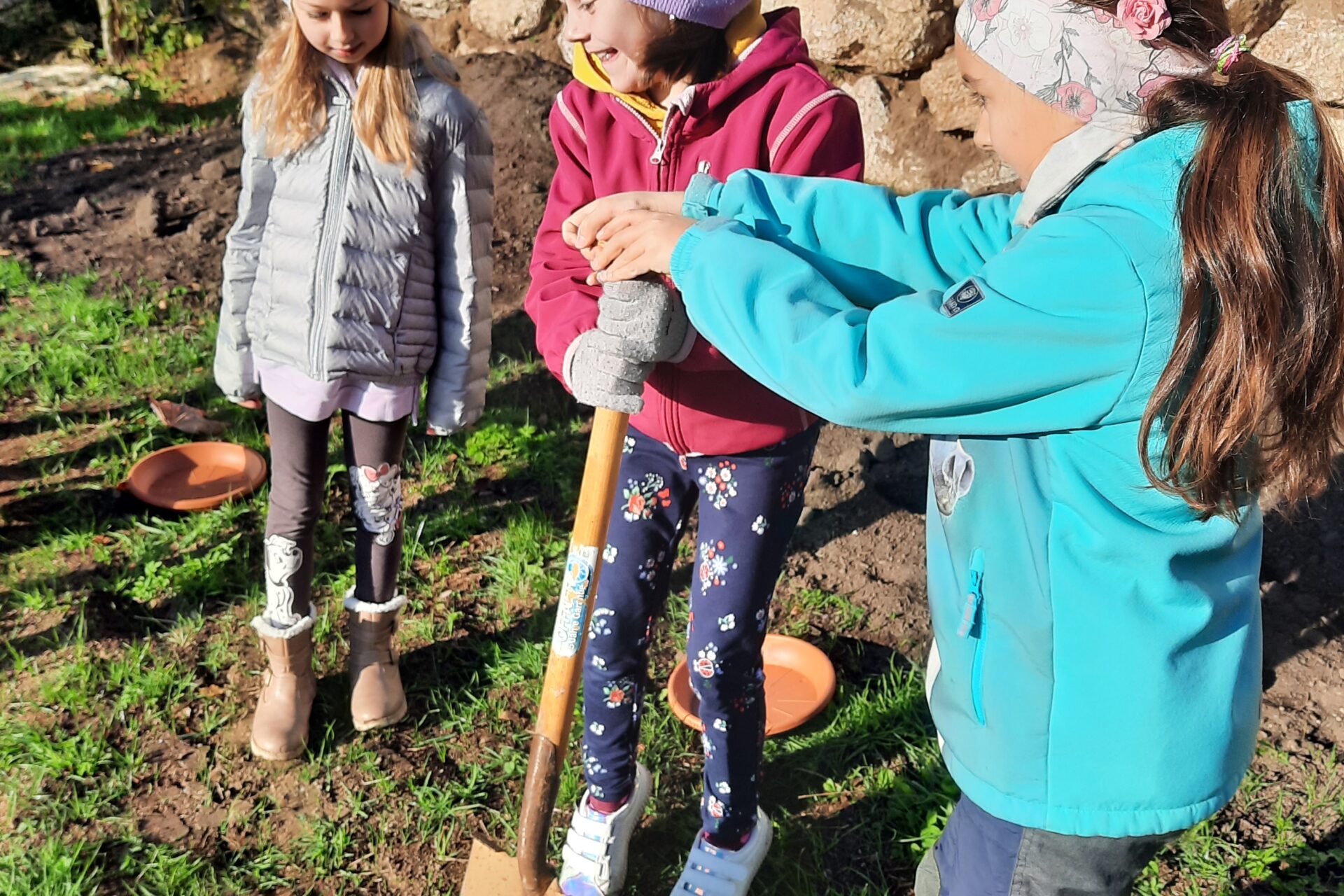 Drei Kinder stehen in Winterkleidung auf einer Wiese und buddeln mit einer Schaufel