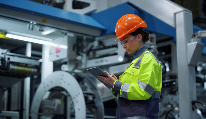 Eine Frau in technischer Arbeitskleidung mit Helm, vertieft in ihr Tablet, in einer Werkshalle.