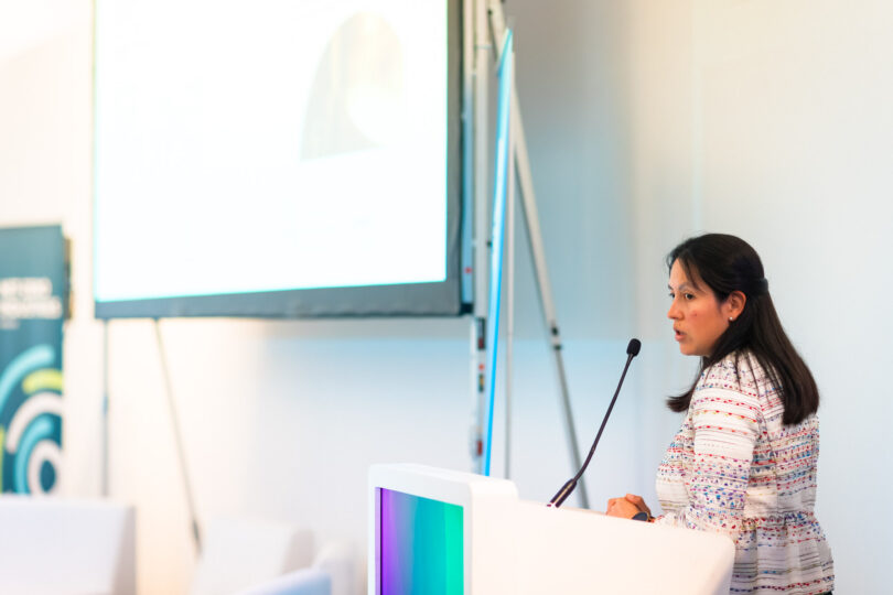Frau mit dunklen Haaren und einem Shirt mit Muster spricht hinter einem Pult. Dahinter ist ein Beamer