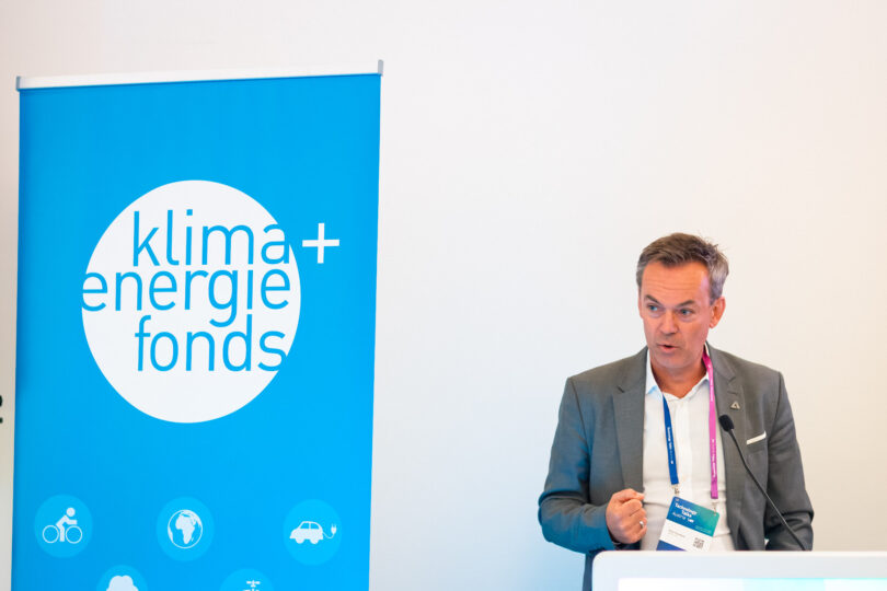 Mann hält eine Rede hinter einem Podium mit Jackett und weißem Hemd. Links neben ihm steht ein Roll Up mit Klimafonds-Logo