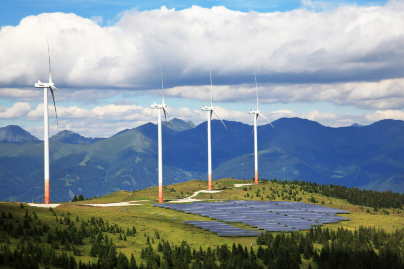 Mehrere Windräder und Solarpaneele auf einem Berghang erzeugen erneuerbare Energie. Im Hintergrund sind bewaldete Berge und Wolken am Himmel zu sehen – Symbol für nachhaltige Energie in der Natur.