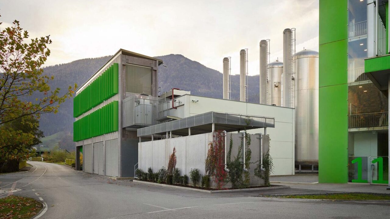 Industrieanlage mit grünen und weißen Gebäudeteilen, mehreren hohen silbernen Tanks und Bergen im Hintergrund.