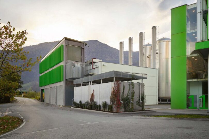 Industrieanlage mit grünen und weißen Gebäudeteilen, mehreren hohen silbernen Tanks und Bergen im Hintergrund.