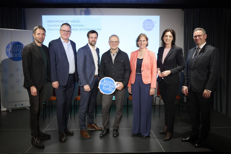 Sieben Personen in Businesskleidung stehen nebeneinander auf einer Bühne, eine Person hält ein rundes Schild mit der Aufschrift 'klima energie fonds'. Im Hintergrund ist eine Präsentationsfolie mit dem Text 'INNOVATION IM DIALOG: Transformation Schiene - Österreichs Sektor als Erfolgsmodell' zu sehen.