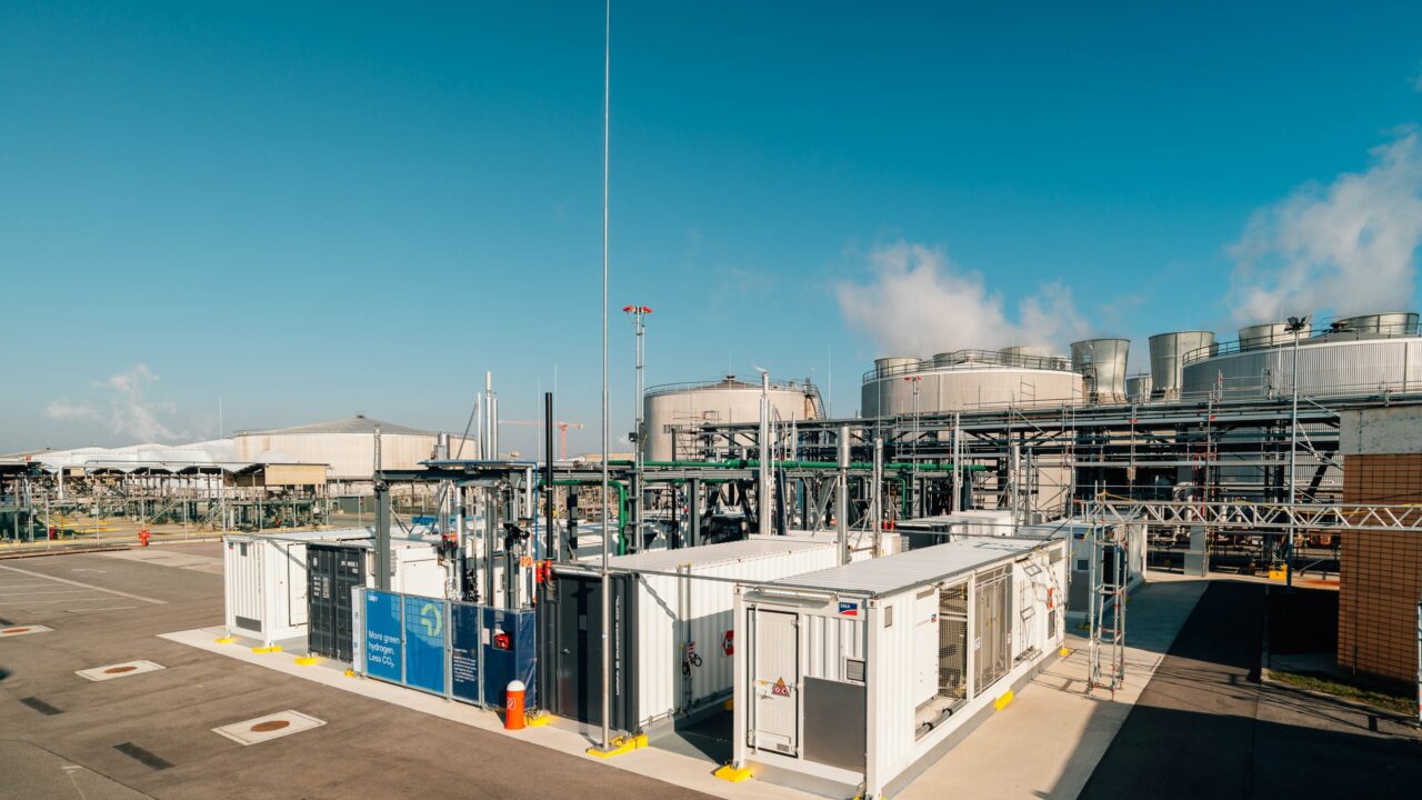 Industrieanlage mit mehreren großen zylindrischen Tanks und weißen Containern auf Asphaltfläche unter blauem Himmel.