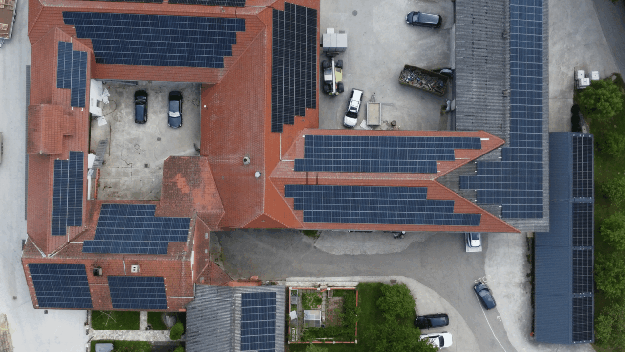 Luftaufnahme eines Gebäudekomplexes mit roten Dächern, auf denen mehrere Solarpaneele installiert sind, umgeben von Parkplätzen und Bäumen.