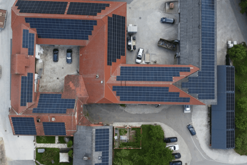 Luftaufnahme eines Gebäudekomplexes mit roten Dächern, auf denen mehrere Solarpaneele installiert sind, umgeben von Parkplätzen und Bäumen.
