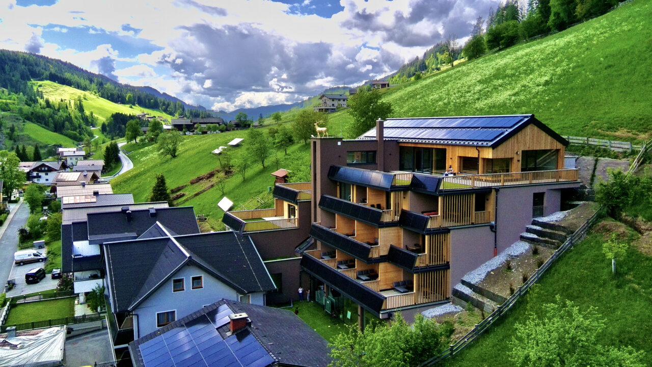 Mehrstöckiges Aparthotel mit Holzfassade und Solarpaneelen auf dem Dach, umgeben von grünen Hügeln und weiteren Gebäuden.