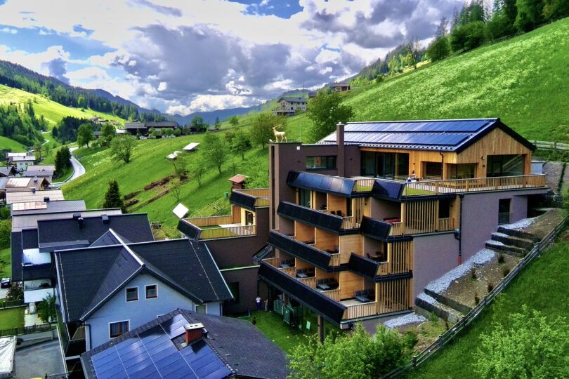 Mehrstöckiges Aparthotel mit Holzfassade und Solarpaneelen auf dem Dach, umgeben von grünen Hügeln und weiteren Gebäuden.