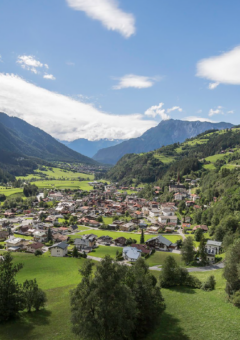 Blick auf ein Tal mit einem Dorf, umgeben von bewaldeten Bergen und grünen Wiesen unter einem bewölkten Himmel.