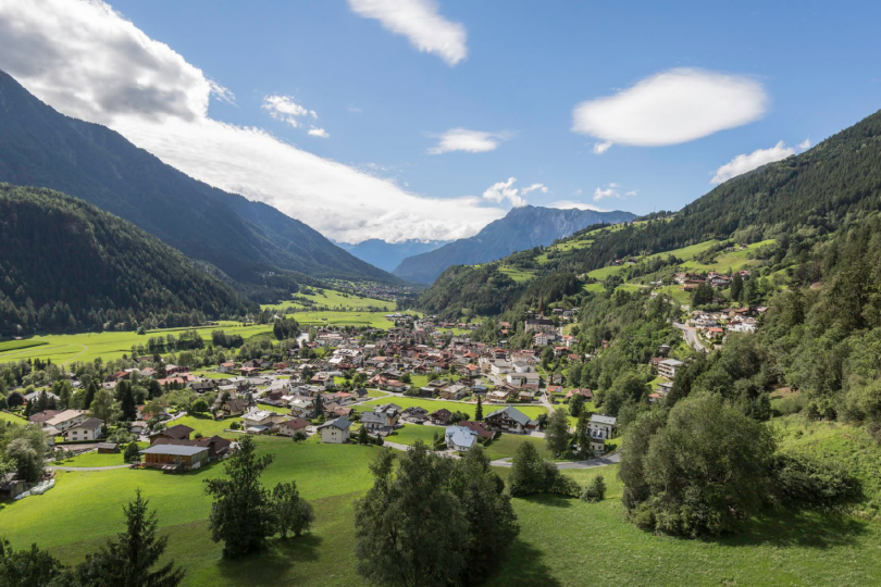 Blick auf ein Tal mit einem Dorf, umgeben von bewaldeten Bergen und grünen Wiesen unter einem bewölkten Himmel.