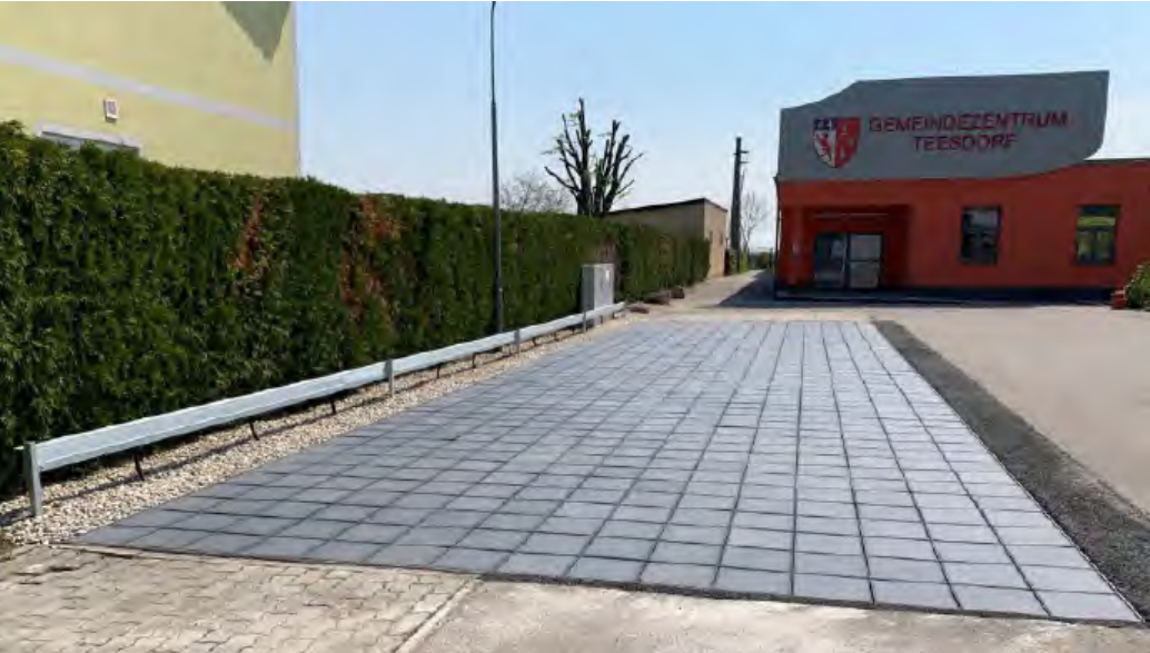 Gebäude mit roter Fassade und Schrift 'GEMEINDEZENTRUM TEESDORF', davor gepflasterter Parkplatz mit einer PV-Anlage am Boden und Hecke entlang eines Gehwegs.