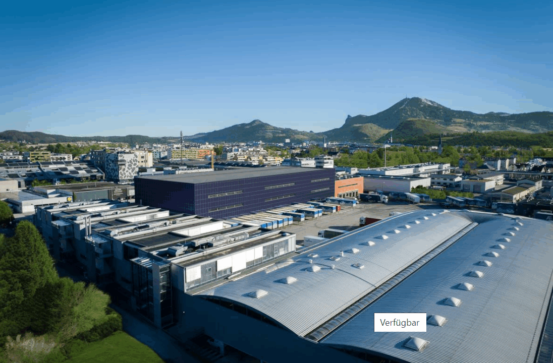 Luftaufnahme eines Industriegebiets mit mehreren großen Hallen und Bergen im Hintergrund unter blauem Himmel.