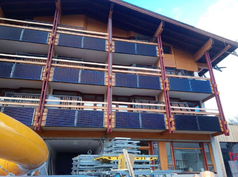 Dreistöckiges Holzgebäude mit Balkonen, darunter Baustellenmaterialien und gelbe Rutsche sichtbar.