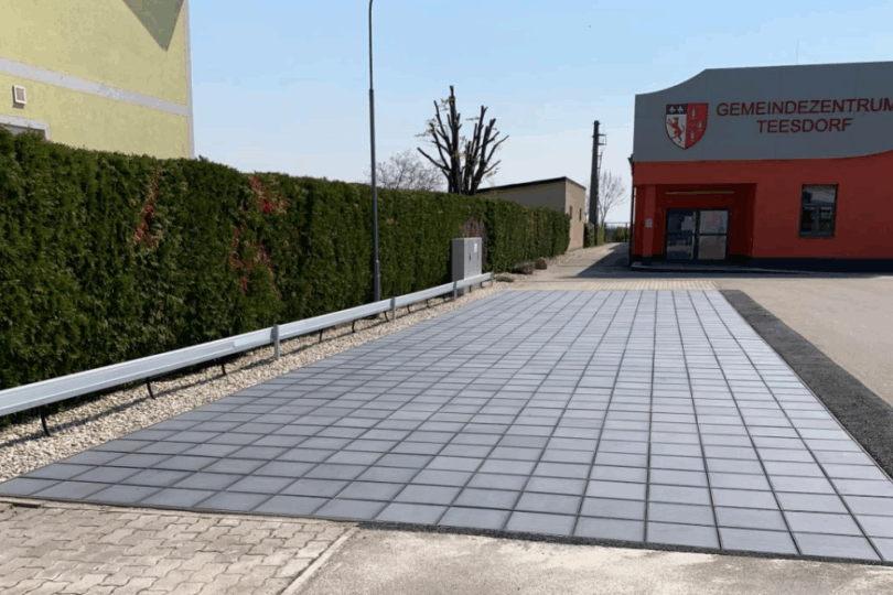 Gebäude mit roter Fassade und Schrift 'GEMEINDEZENTRUM TEESDORF', davor gepflasterter Parkplatz mit einer PV-Anlage am Boden und Hecke entlang eines Gehwegs.