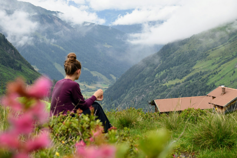 Frau mit hochgestecktem Haar sitzt auf einer Wiese und blickt auf ein Tal mit Bergen und einem Haus mit rotem Dach.