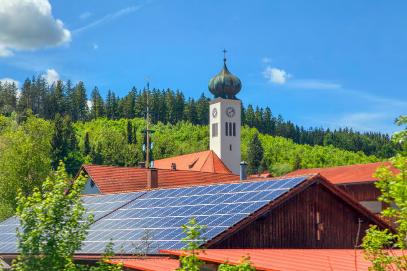 Dach mit Solarpaneelen vor bewaldetem Hügel und Kirchturm mit Zwiebelturm unter blauem Himmel