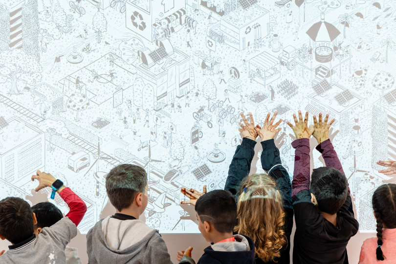 Man sieht Kinder von hinten, die vor einer Wand stehen, auf die eine grafisch gestaltete Darstellung einer Stadt der Zukunft projiziert wird.