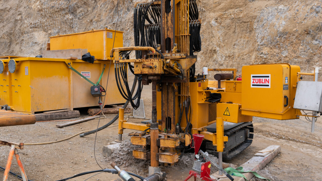Man sieht ein gelbes Tiefenbohrgerät auf einer Baustelle umgeben von Felsen.