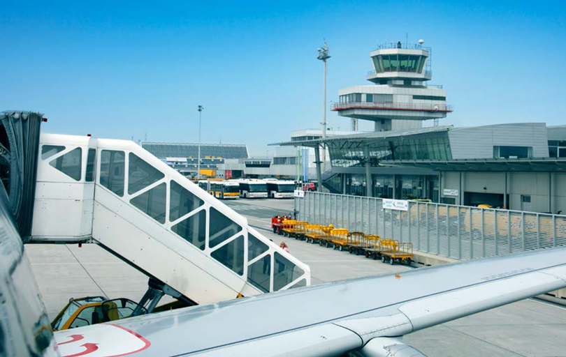 Ein Flughafen im Hintergrund, im Vordergrund ein Flugzeugflügel und die elektrische Treppe zum Einstieg.