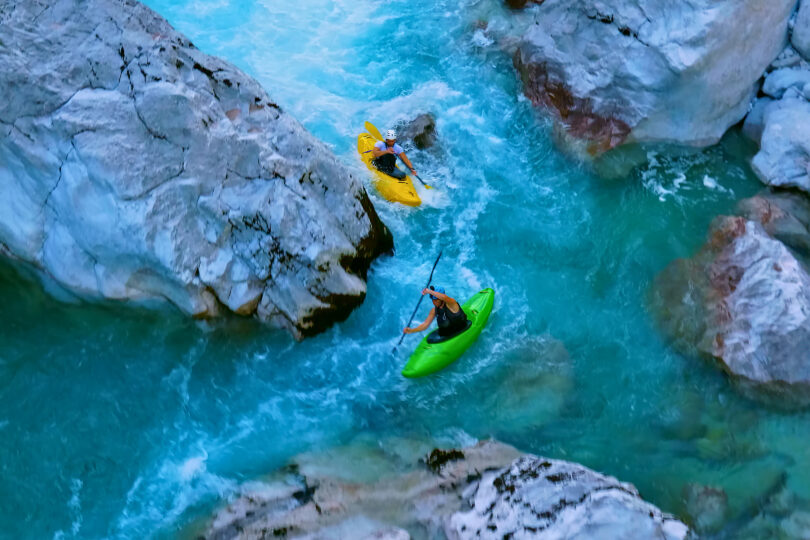 Foto: Eine atemberaubende Luftaufnahme zeigt zwei Kajakfahrer, die einen schmalen, reißenden Fluss zwischen großen Felsen hinunterfahren. Ein Kajakfahrer mit weißem Helm steuert ein gelbes Kajak, während der andere, in schwarzer Kleidung, ein grünes Kajak manövriert. Die Szene verdeutlicht den Nervenkitzel und die Herausforderung des Extrem-Kajakfahrens in einer engen, felsigen Schlucht.